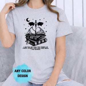 May include: Light gray t-shirt featuring a black graphic of stacked books, a crescent moon, stars, and a dragon. The text "I AM THE SKY AND THE POWER OF EVERY STORM THAT HAS EVER BEEN. I AM INFINITE" is below the books. "ANY COLOR DESIGN" is at the bottom.