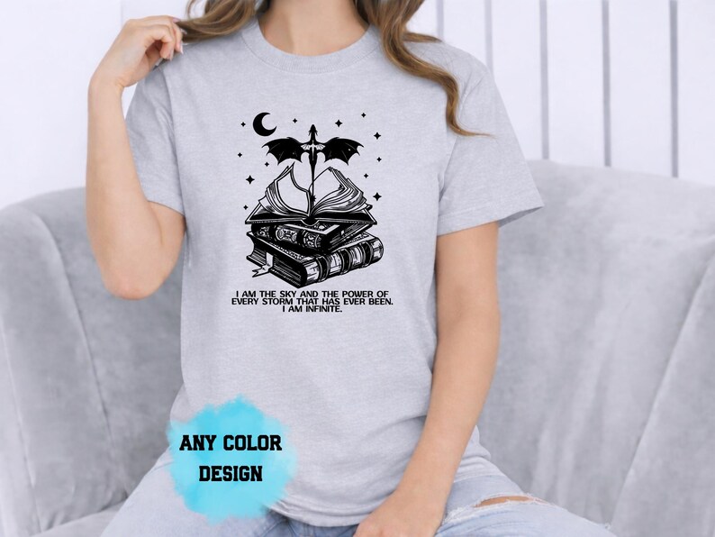 May include: Light gray t-shirt featuring a black graphic of stacked books, a crescent moon, stars, and a dragon. The text "I AM THE SKY AND THE POWER OF EVERY STORM THAT HAS EVER BEEN. I AM INFINITE" is below the books. "ANY COLOR DESIGN" is at the bottom.