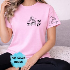 May include: A light pink t-shirt featuring a black graphic of roller skates. The sleeve has a smaller roller skate graphic with the name "Jennifer" in cursive. The shirt also has the text "ANY COLOR DESIGN".