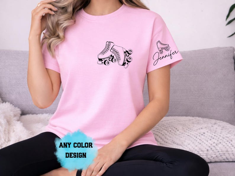 May include: A light pink t-shirt featuring a black graphic of roller skates. The sleeve has a smaller roller skate graphic with the name "Jennifer" in cursive. The shirt also has the text "ANY COLOR DESIGN".