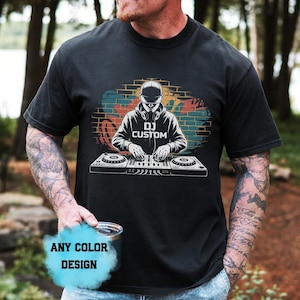 May include: A black short-sleeved t-shirt featuring a graphic print of a DJ wearing a cap and headphones, positioned behind a mixing console. The background of the graphic shows a brick wall design. The text "DJ CUSTOM" is printed on the shirt.