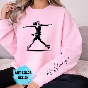 Puede incluir: Sudadera rosa claro con un gráfico negro de una patinadora artística en el aire, con los brazos extendidos. La manga derecha tiene el nombre "Jennifer" en cursiva y una pequeña ilustración de patines de hielo. El texto "ANY COLOR DESIGN" está en la parte inferior.