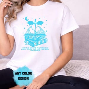 May include: White t-shirt with a light blue graphic design. The design features a stack of books, a crescent moon, stars, and text that reads "I AM THE SKY AND THE POWER OF EVERY STORM THAT HAS EVER BEEN. I AM INFINITE." The words "ANY COLOR DESIGN" are also visible.