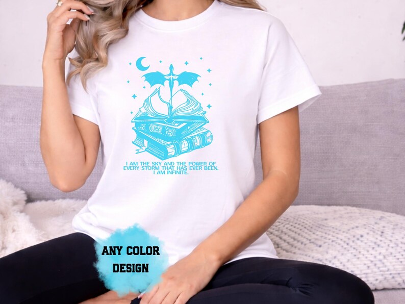 May include: White t-shirt with a light blue graphic design. The design features a stack of books, a crescent moon, stars, and text that reads "I AM THE SKY AND THE POWER OF EVERY STORM THAT HAS EVER BEEN. I AM INFINITE." The words "ANY COLOR DESIGN" are also visible.