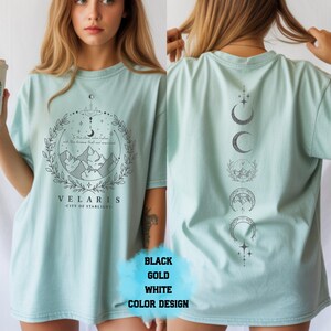 Pode incluir: Camiseta azul claro com um gr&aacute;fico frontal com montanhas, uma lua crescente e o texto "VELARIS CITY OF STARLIGHT". A parte de tr&aacute;s da camisa exibe uma s&eacute;rie de fases da lua. A camisa est&aacute; dispon&iacute;vel em preto, dourado e branco.