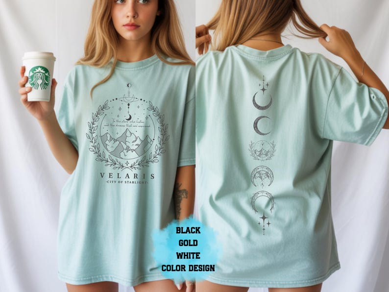 Pode incluir: Camiseta azul claro com um gr&aacute;fico frontal com montanhas, uma lua crescente e o texto "VELARIS CITY OF STARLIGHT". A parte de tr&aacute;s da camisa exibe uma s&eacute;rie de fases da lua. A camisa est&aacute; dispon&iacute;vel em preto, dourado e branco.