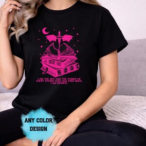 May include: Black t-shirt with a pink graphic featuring a stack of books, a crescent moon, and a dragon. The text "I AM THE SKY AND THE POWER OF EVERY STORM THAT HAS EVER BEEN. I AM INFINITE" is printed below. The design is available in any color.