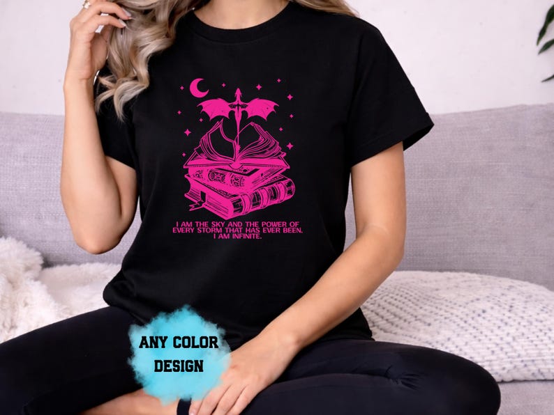 May include: Black t-shirt with a pink graphic featuring a stack of books, a crescent moon, and a dragon. The text "I AM THE SKY AND THE POWER OF EVERY STORM THAT HAS EVER BEEN. I AM INFINITE" is printed below. The design is available in any color.