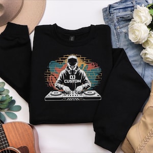 May include: A black long-sleeve sweatshirt featuring a graphic print of a DJ wearing a cap and headphones, positioned behind a mixing console. The background of the graphic shows a brick wall with colourful graffiti accents and the text DJ CUSTOM.