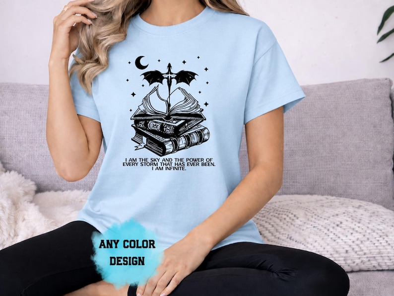 May include: Light blue t-shirt with a black graphic of stacked books, a sword, moon, and stars. Text on the shirt reads "I AM THE SKY AND THE POWER OF EVERY STORM THAT HAS EVER BEEN. I AM INFINITE." The words "ANY COLOR DESIGN" are also visible.