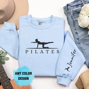 Könnte beinhalten: Ein hellblaues Sweatshirt mit Rundhalsausschnitt und einem schwarzen Aufdruck einer Person in Pilates-Pose über dem Schriftzug PILATES. Der linke Ärmel zeigt den Namen Jennifer in schwarzer Schrift. Ein blauer Kreis enthält den Text ANY COLOR DESIGN.