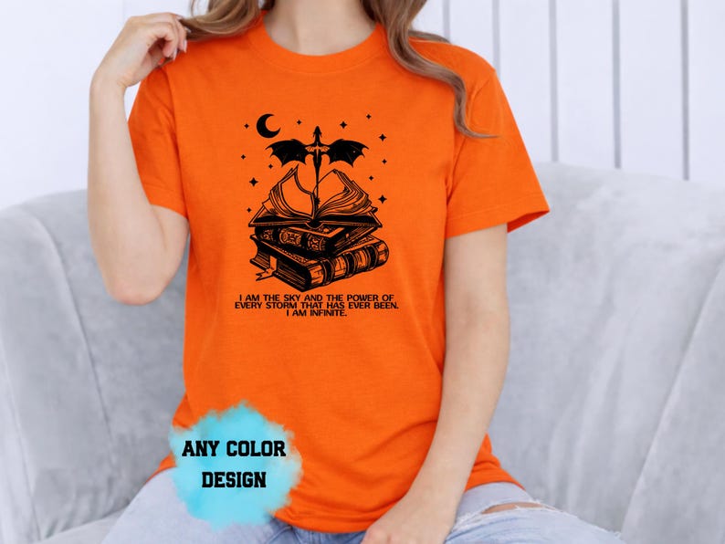 May include: Orange t-shirt with a black graphic of stacked books, a dragon, moon, and stars. Text on the shirt reads "I AM THE SKY AND THE POWER OF EVERY STORM THAT HAS EVER BEEN. I AM INFINITE." The shirt design is available in any color.