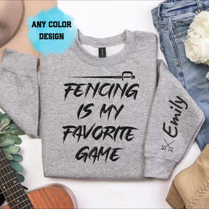 May include: A heather gray crewneck sweatshirt featuring black text that reads FENCING IS MY FAVORITE GAME with a sword icon above it. The left sleeve displays the name Emily in black script above a small crossed swords graphic.