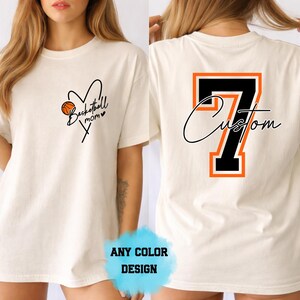 May include: Cream-colored t-shirt with a basketball-themed design. The front features a heart with a basketball and the words "Basketball Mom." The back displays a large number 7 with "Custom" written across it. The shirt is a casual style.