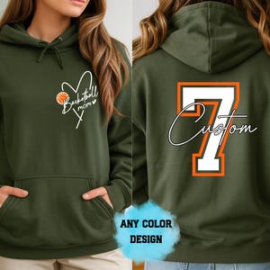Può includere: Felpa con cappuccio verde oliva con design a tema basket. La parte anteriore presenta un cuore con un pallone da basket e la scritta "Basketball mom". Il retro mostra il numero 7 con "Custom" in corsivo.
