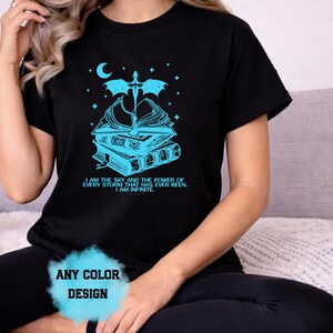 May include: Black t-shirt with a teal graphic of books, a sword, and a crescent moon. The text reads "I AM THE SKY AND THE POWER OF EVERY STORM THAT HAS EVER BEEN. I AM INFINITE." The words "ANY COLOR DESIGN" are also visible.