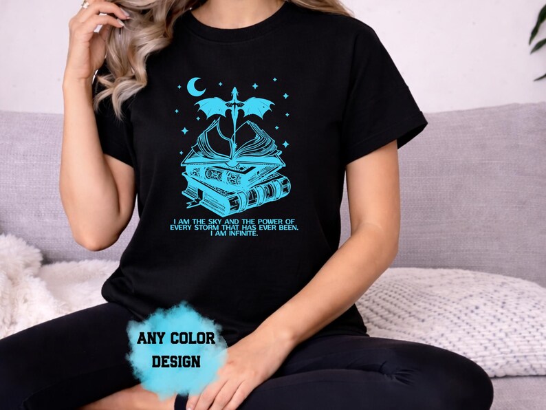 May include: Black t-shirt with a teal graphic of books, a sword, and a crescent moon. The text reads "I AM THE SKY AND THE POWER OF EVERY STORM THAT HAS EVER BEEN. I AM INFINITE." The words "ANY COLOR DESIGN" are also visible.