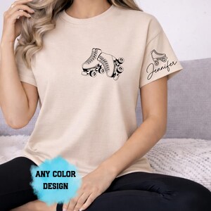May include: Beige t-shirt featuring a black graphic of two roller skates on the front. The right sleeve has a smaller roller skate graphic with the name "Jennifer" in script. The bottom of the shirt has the text "ANY COLOR DESIGN".