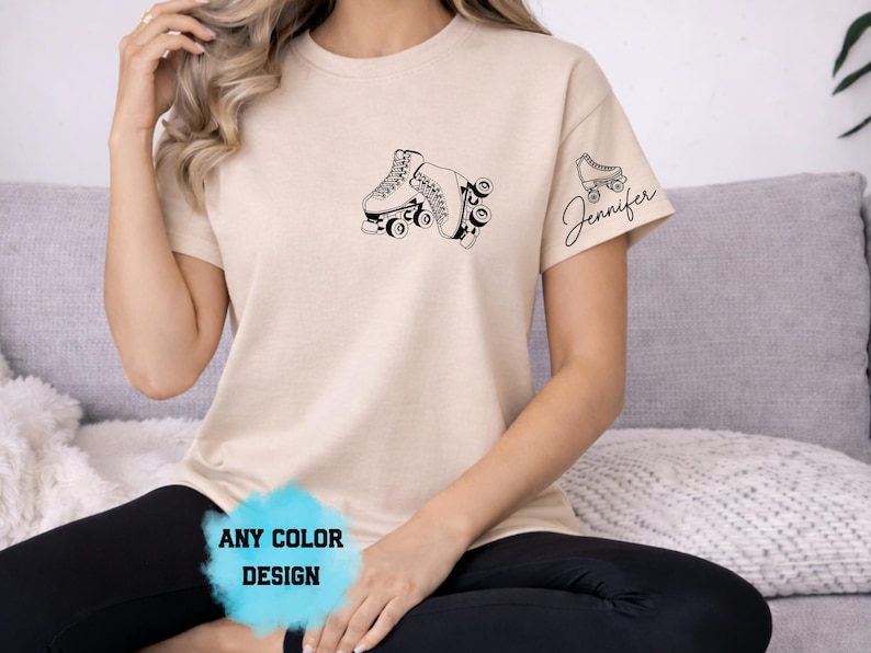May include: Beige t-shirt featuring a black graphic of two roller skates on the front. The right sleeve has a smaller roller skate graphic with the name "Jennifer" in script. The bottom of the shirt has the text "ANY COLOR DESIGN".