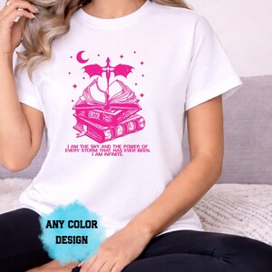 May include: White t-shirt with a pink graphic design featuring a stack of books, a sword, a crescent moon, and stars. The text "I AM THE SKY AND THE POWER OF EVERY STORM THAT HAS EVER BEEN. I AM INFINITE." is printed below the graphic. The words "ANY COLOR DESIGN" are also visible.