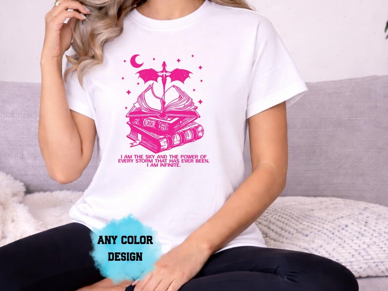 May include: White t-shirt with a pink graphic design featuring a stack of books, a sword, a crescent moon, and stars. The text "I AM THE SKY AND THE POWER OF EVERY STORM THAT HAS EVER BEEN. I AM INFINITE." is printed below the graphic. The words "ANY COLOR DESIGN" are also visible.