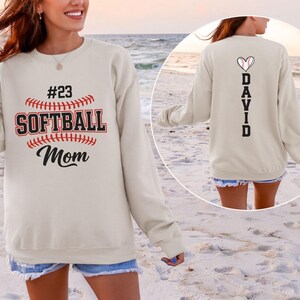 Puede incluir: Una sudadera beige de cuello redondo con un gráfico en la parte delantera que incluye el texto #23 SOFTBALL Mom y una ilustración de béisbol. La parte trasera muestra el nombre DAVID en vertical con un icono de corazón de béisbol.