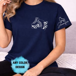 May include: Navy blue t-shirt with a white roller skate graphic on the front and a smaller roller skate design with the name "Jennifer" on the sleeve. The shirt has a crew neck and short sleeves. The text "ANY COLOR DESIGN" is also visible.