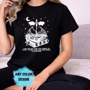 May include: Black t-shirt with a white graphic featuring a stack of books, a sword, a crescent moon, and stars. The text reads "I AM THE SKY AND THE POWER OF EVERY STORM THAT HAS EVER BEEN. I AM INFINITE." The design is available in any color.