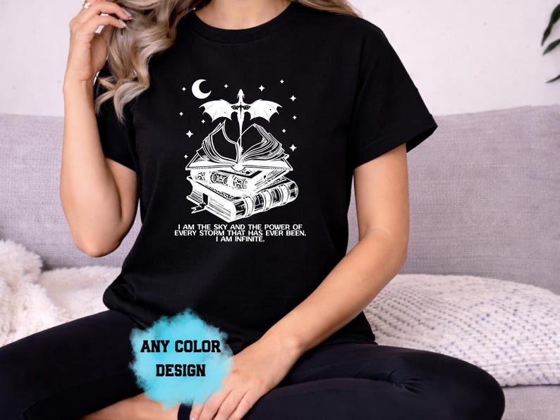 May include: Black t-shirt with a white graphic featuring a stack of books, a sword, a crescent moon, and stars. The text reads "I AM THE SKY AND THE POWER OF EVERY STORM THAT HAS EVER BEEN. I AM INFINITE." The design is available in any color.