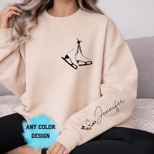 May include: Beige sweatshirt with a black graphic of ice skates. The text "Jennifer" is written in cursive on the sleeve. The words "ANY COLOR DESIGN" are in a blue box at the bottom.