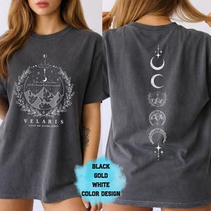 Pode incluir: T-shirt cinza escuro com um gr&aacute;fico branco na frente com montanhas, estrelas e o texto "VELARIS - CITY OF STARLIGHT". A parte de tr&aacute;s da camisa tem um design vertical de fases da lua. A camisa &eacute; um estilo unissex.