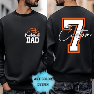 May include: A black crewneck sweatshirt featuring a basketball graphic and the text Basketball DAD on the front left chest. The back displays a large white number 7 with an orange outline and the word Custom in white script text.
