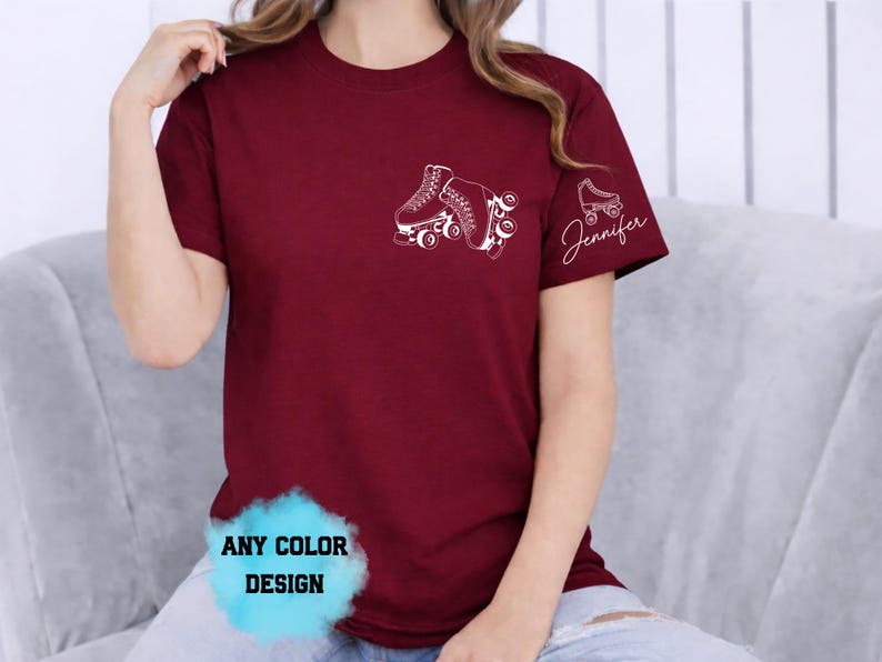 May include: Burgundy t-shirt with a white roller skate graphic on the chest and sleeve. The sleeve also features the name "Jennifer" in script. The shirt has a crew neck and short sleeves. The bottom of the shirt has a blue and white graphic that says "ANY COLOR DESIGN".