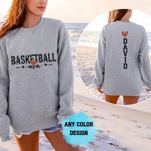 Puede incluir: Una sudadera gris jaspeado de manga larga con el texto negro "BASKETBALL mom" y un corazón de baloncesto naranja en el frente, y el texto vertical negro "DAVID" con un corazón de baloncesto naranja encima en la parte posterior.