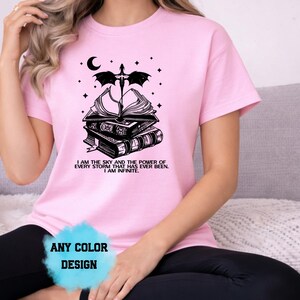 May include: Pink t-shirt with a black graphic of stacked books, a crescent moon, stars, and a dragon. Text on the shirt reads "I AM THE SKY AND THE POWER OF EVERY STORM THAT HAS EVER BEEN. I AM INFINITE." The shirt design is available in any color.