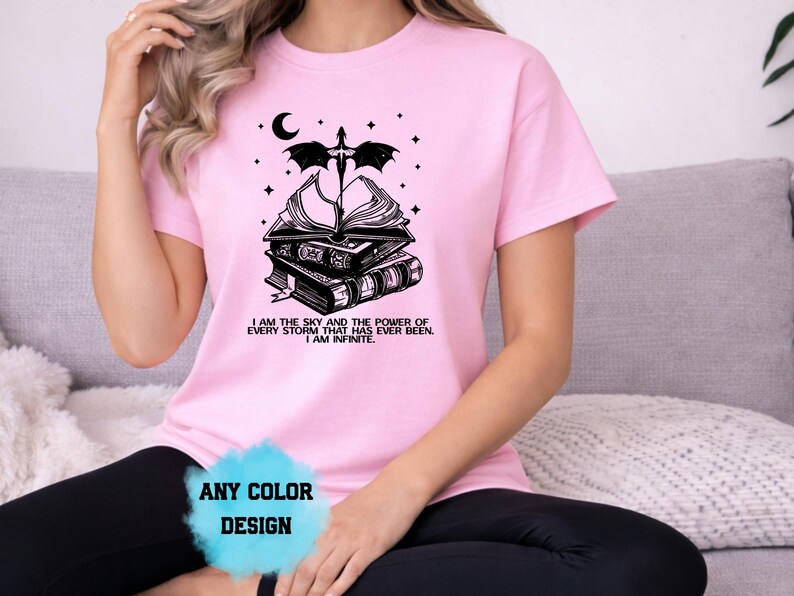 May include: Pink t-shirt with a black graphic of stacked books, a crescent moon, stars, and a dragon. Text on the shirt reads "I AM THE SKY AND THE POWER OF EVERY STORM THAT HAS EVER BEEN. I AM INFINITE." The shirt design is available in any color.