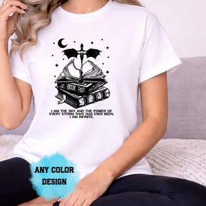 May include: White t-shirt with a black graphic design featuring a stack of books, a dragon, a crescent moon, and stars. The text "I AM THE SKY AND THE POWER OF EVERY STORM THAT HAS EVER BEEN. I AM INFINITE." is printed below the design. The words "ANY COLOR DESIGN" are also visible.