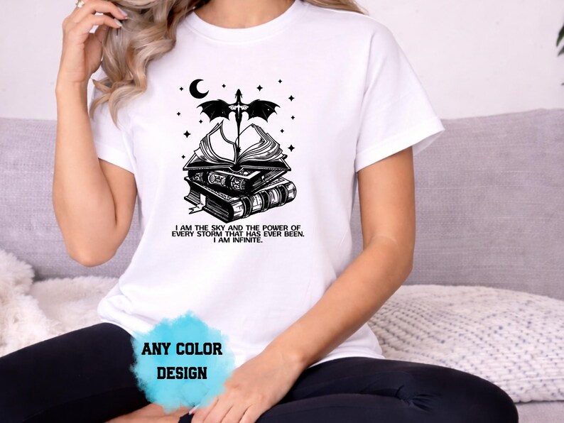 May include: White t-shirt with a black graphic design featuring a stack of books, a dragon, a crescent moon, and stars. The text "I AM THE SKY AND THE POWER OF EVERY STORM THAT HAS EVER BEEN. I AM INFINITE." is printed below the design. The words "ANY COLOR DESIGN" are also visible.