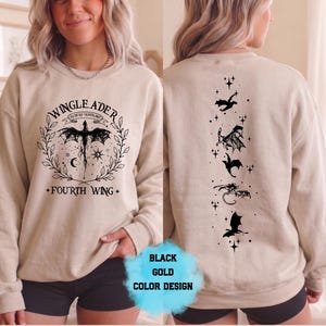 May include: Beige sweatshirt with black dragon graphic and text "WINGLEADER" and "FOURTH WING". The back features a vertical line of black dragon silhouettes and stars. A "BLACK GOLD COLOR DESIGN" label is visible.