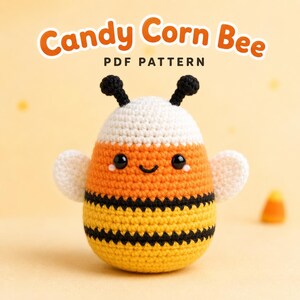 May include: A crocheted bee toy with a candy corn color scheme of white, orange, and yellow stripes with black accents. The bee has two black antennae, small white wings, and a smiling face. The text Candy Corn Bee PDF PATTERN is above.