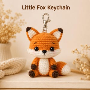 May include: A crochet fox keychain featuring the text Little Fox Keychain. The fox is orange with a white face, chest, and tail tip, and dark brown ears and paws. It has black plastic eyes and a small black nose, attached to a silver metal clasp.