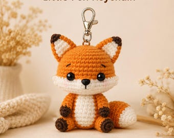 May include: A crochet fox keyring featuring the text Little Fox Keychain. The fox is orange with a white face, chest, and tail tip, and dark brown ears and paws. It has black plastic eyes and a small black nose, attached to a silver metal clasp.
