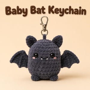 May include: A dark gray crocheted baby bat keychain with black plastic eyes, pink embroidered cheeks, and two small white fangs. The bat has outstretched wings and is attached to a silver metal lobster clasp. The text Baby Bat Keychain is above.