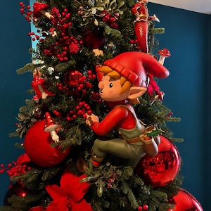 May include: A decorated Christmas tree with red ornaments, berries, and a figurine of a red-clad elf climbing the branches. The tree is adorned with red flowers and mushroom-shaped ornaments, creating a festive holiday display.