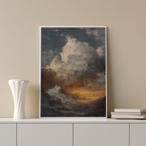 May include: Framed art print featuring a dramatic cloudscape with a sunset. The artwork displays a blend of dark blues, browns, and golden hues, creating a moody and atmospheric scene. The print is in a white frame and is displayed on a white cabinet.