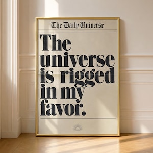 Könnte beinhalten: Ein gerahmtes vertikales Poster mit cremefarbenem Hintergrund und schwarzem Text. Oben steht The Daily Universe. Darunter steht in großer, fetter Schrift The universe is rigged in my favor. Ein kleines Sonnensymbol ist unten zentriert.