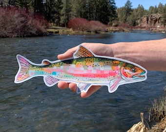 May include: A die-cut sticker featuring a colorful illustration of a rainbow trout. The fish has a vibrant body with pink, orange, and yellow hues, accented by small black spots and a white border. The artwork displays a mosaic-like pattern.