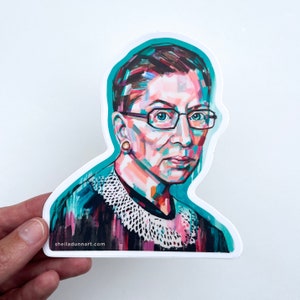 May include: A sticker featuring a colorful portrait of Ruth Bader Ginsburg, a Supreme Court Justice. The sticker has a teal background and the text "sheilladunart.com" at the bottom.