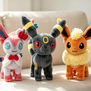 May include: Three plush Pok&eacute;mon figures: Sylveon, Umbreon, and Flareon. Sylveon is pink, white, and blue with a bow. Umbreon is black with yellow accents and red eyes. Flareon is orange with a yellow fur collar. All are standing on a beige surface.