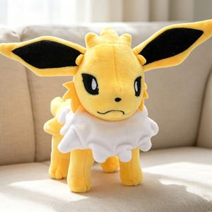 May include: A plush toy of a yellow creature with large, black-tipped ears, black eyes, and a white ruffled collar. The toy is standing on four legs and has a slightly grumpy expression. The toy is a popular character from a well-known franchise.